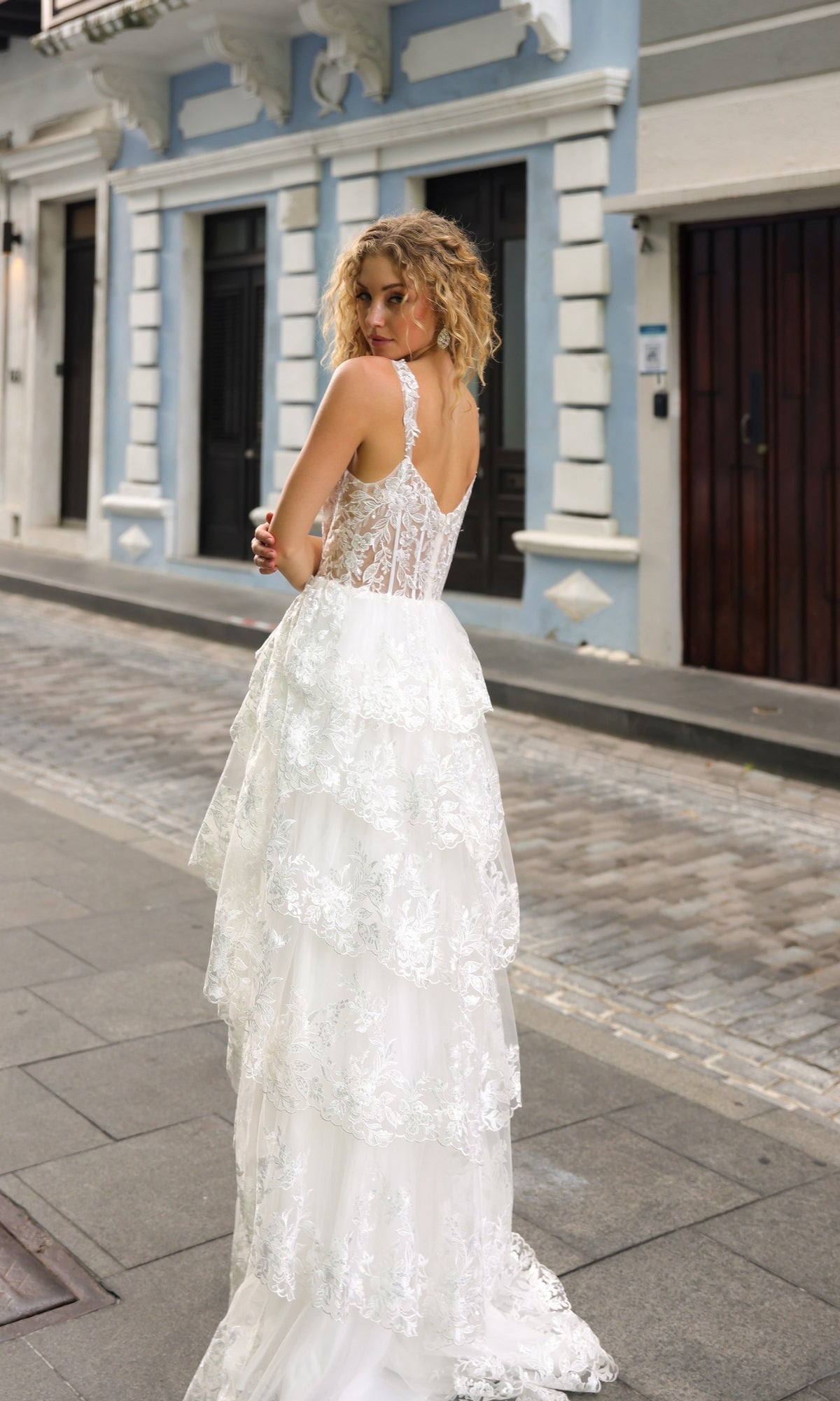 A woman with curly hair stands on a cobblestone street, looking over her shoulder in the Long Lace Ruffled A-Line Prom Dress T1335—perfect for formal events.